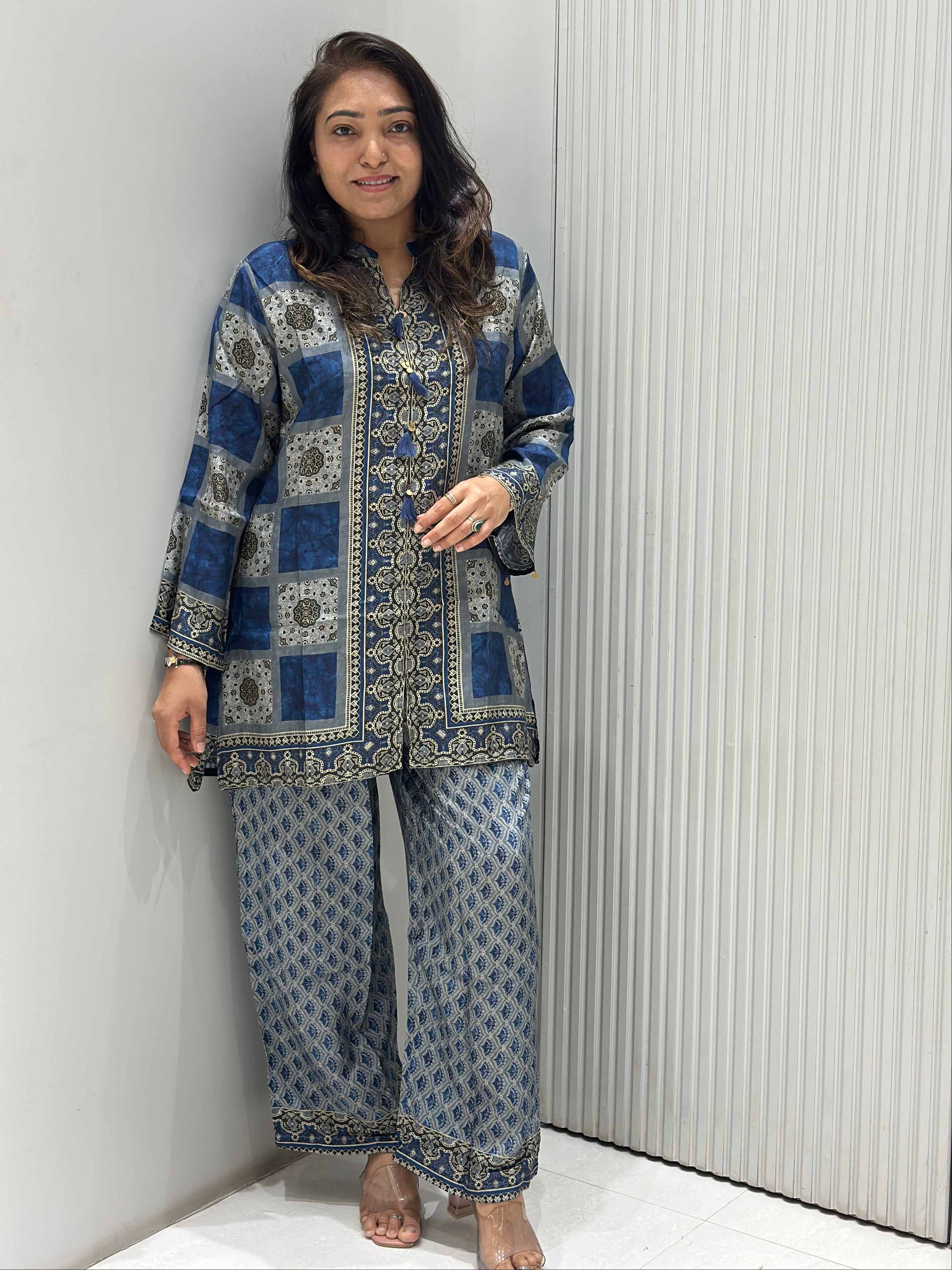A woman wearing a blue and gray patterned traditional kurta set with matching pants, lying down on a white surface against a white slatted wall.
